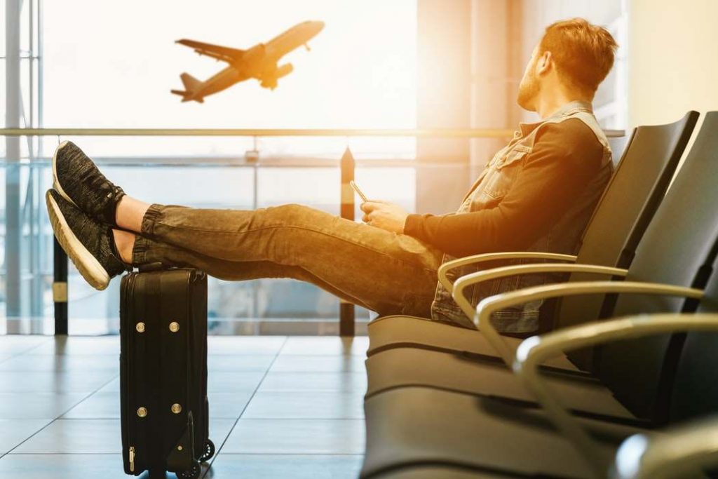 Homem sentado com os pés na mala observando avião voar.