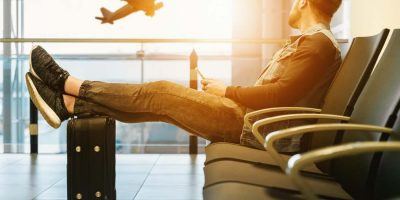 Homem sentado com os pés na mala observando avião voar.