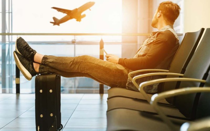 Homem sentado com os pés na mala observando avião voar.